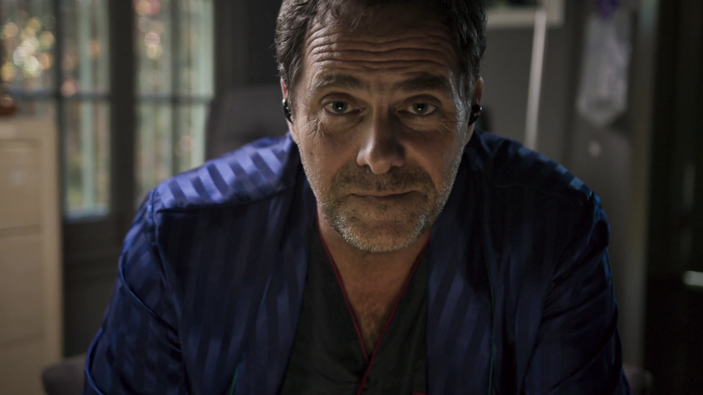 A scruffy man in a blue robe leans towards the webcam