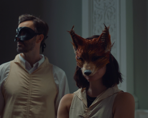 Two people are standing in a hallway. To the left and slightly behind is a brown-haired man wearing a white dress shirt, beige robe, and a black mask covering his eyes and nose. To the right, and slightly to the front, is a brown-haired woman with a black tank top, a beige robe, and a fox mask covering her eyes and nose.