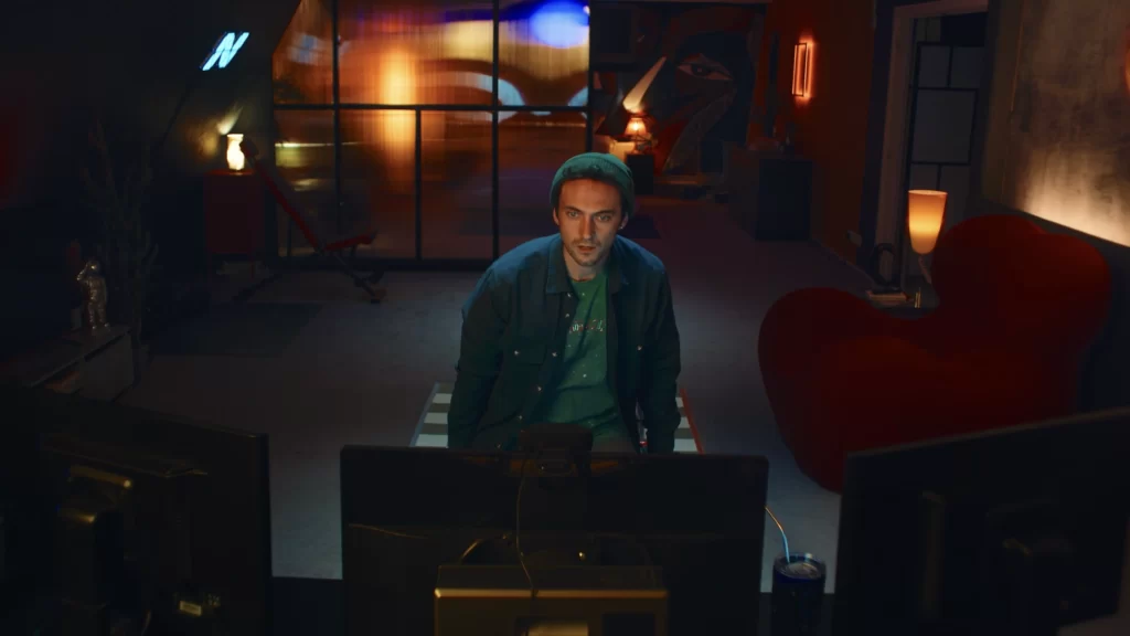 A screenshot from Hello Stranger. Taken from behind a computer monitor, it shows Cam, a man with short hair and a green toque, looking at the monitor. You can see his apartment behind him.