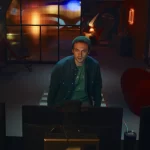 A screenshot from Hello Stranger. Taken from behind a computer monitor, it shows Cam, a man with short hair and a green toque, looking at the monitor. You can see his apartment behind him.