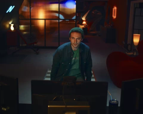 A screenshot from Hello Stranger. Taken from behind a computer monitor, it shows Cam, a man with short hair and a green toque, looking at the monitor. You can see his apartment behind him.