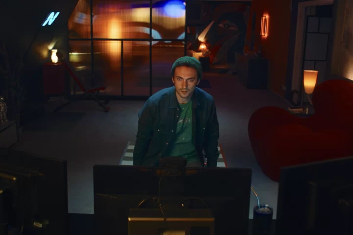 A screenshot from Hello Stranger. Taken from behind a computer monitor, it shows Cam, a man with short hair and a green toque, looking at the monitor. You can see his apartment behind him.