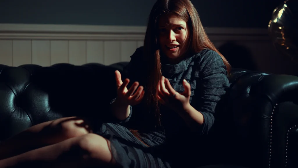 A screenshot of Ophelia in The Infectious Madness of Doctor Dekker 2. She is a girl with long brown hair, sitting on a dark couch. She is wearing all black, and she's holding up her cupped hands in front of her body.